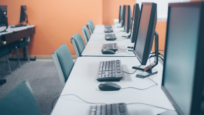 Telpa ar rindā izvietotiem galddatoriem, monitoriem, tastatūrām un pelēm uz balta galda pie oranžas sienas.