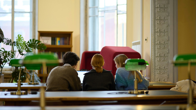 Attēls ar bibliotēku, kurā pie datora sēž vairāki cilvēki palīdzot viens otram