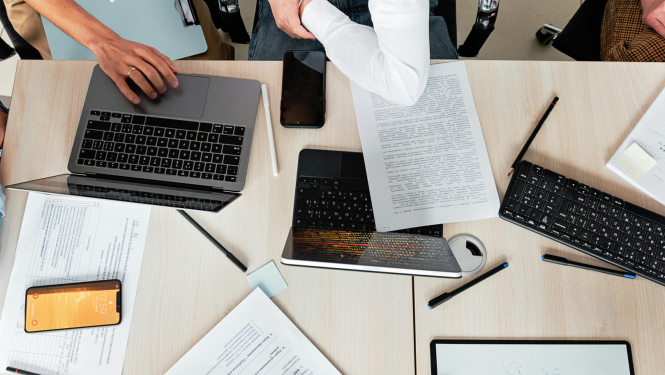 Foto ar cilvēkiem pie galda, uz kura ir vairāki datori, planšetes un dokumenti.