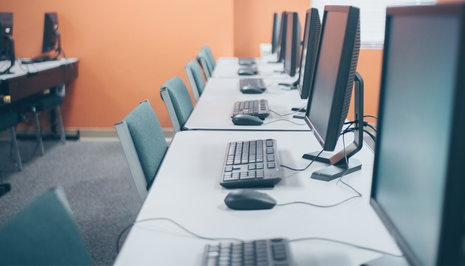 Telpa ar rindā izvietotiem galddatoriem, monitoriem, tastatūrām un pelēm uz balta galda pie oranžas sienas.