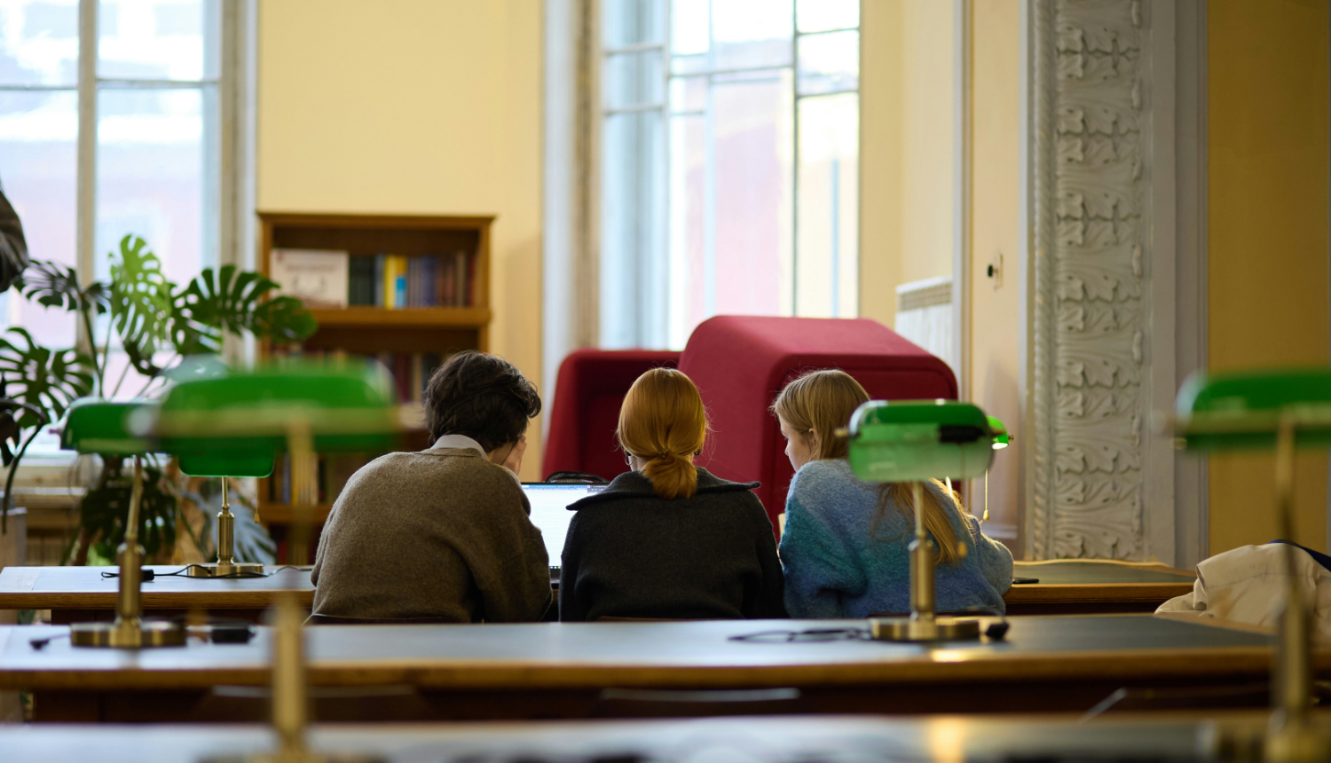 Attēls ar bibliotēku, kurā pie datora sēž vairāki cilvēki palīdzot viens otram